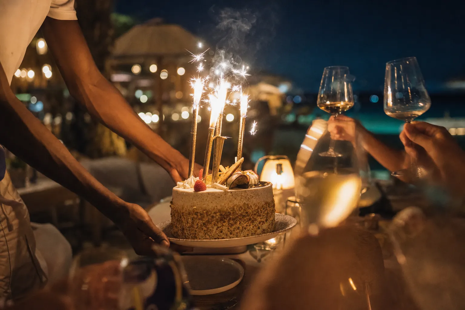 Celebration cake and sparklers arriving at a beach club table at night, guests raising glasses out of focus