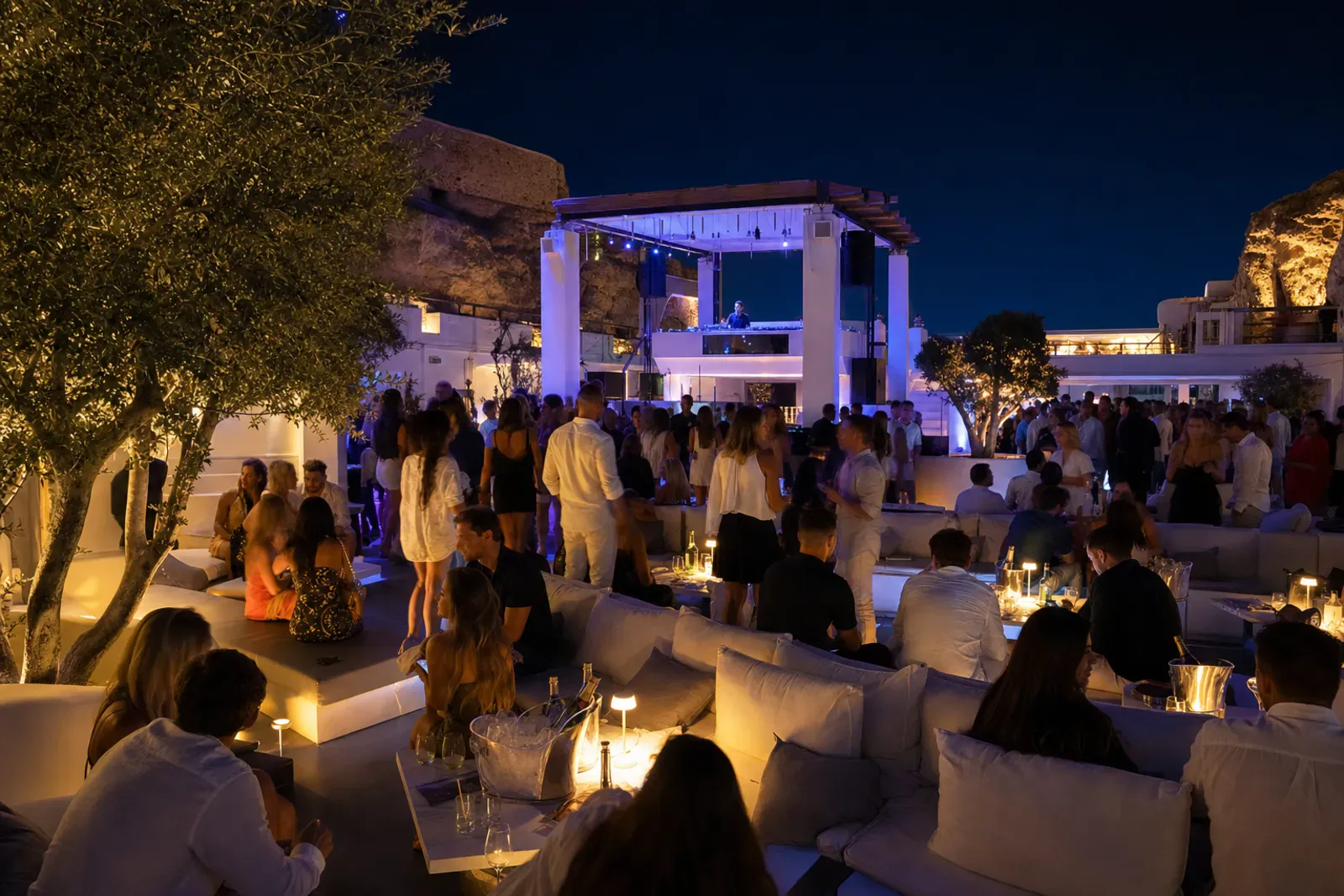 Open-air Mediterranean nightclub at night with VIP tables, low sofas, and a distant DJ booth