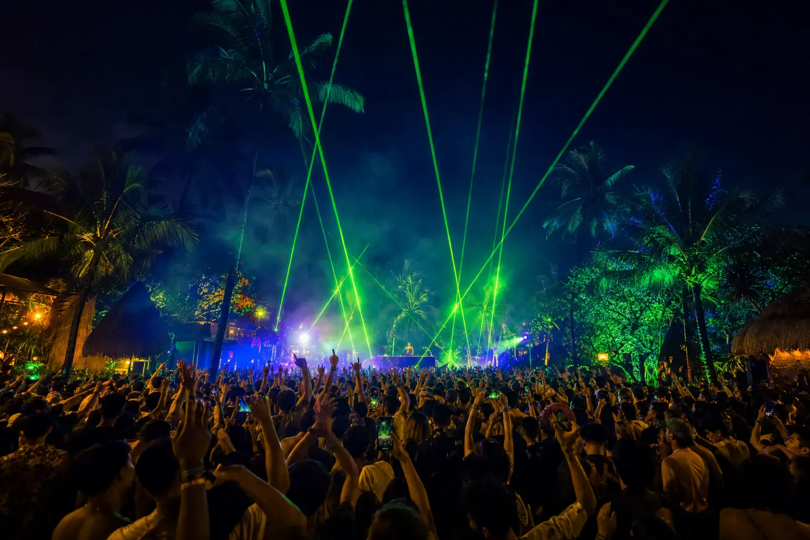Tropical open-air festival crowd at night with green lasers cutting through haze over palm trees