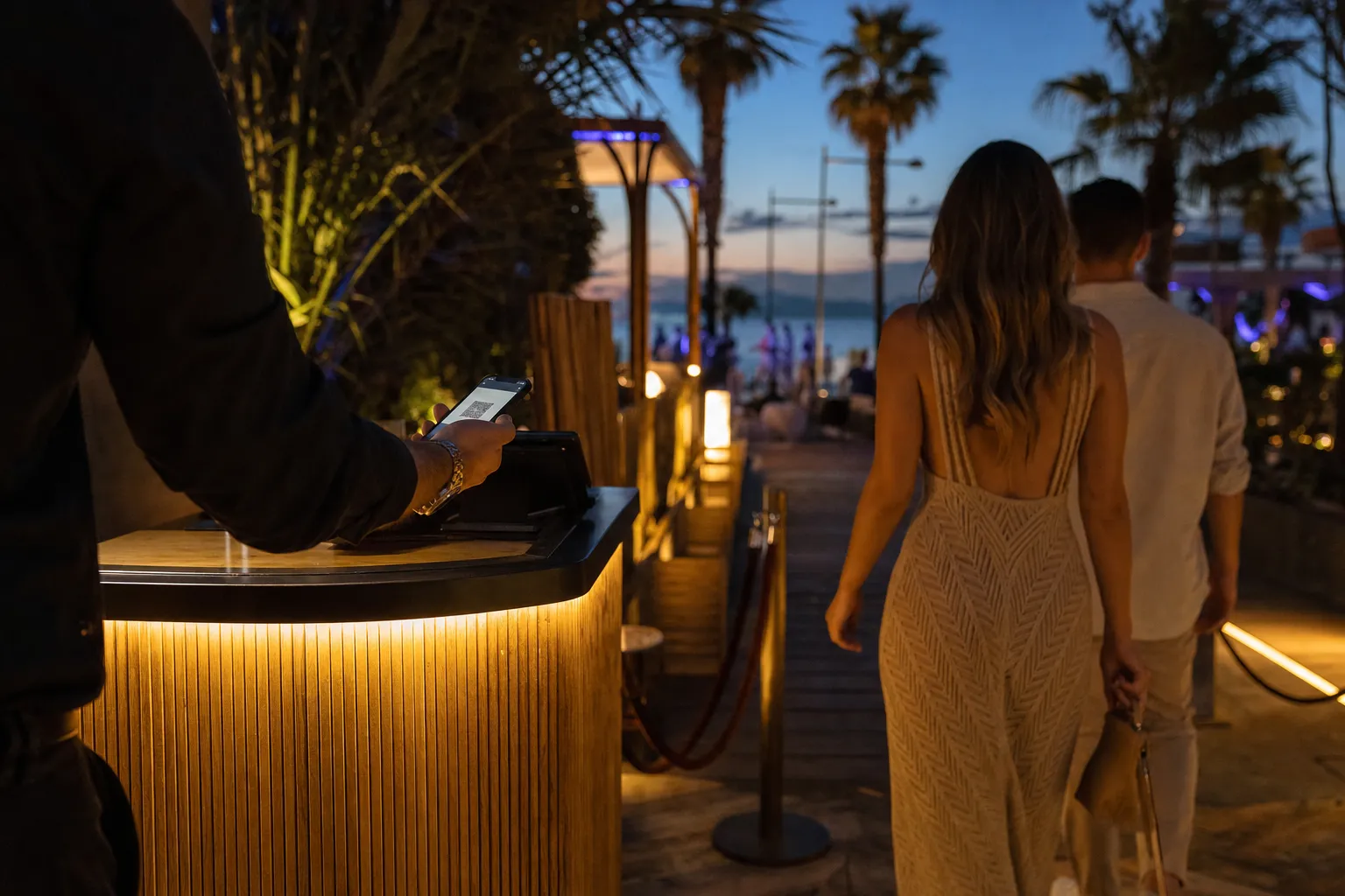 Guests arriving at a beach club entrance at dusk, a staff member scanning a phone at a lit welcome desk