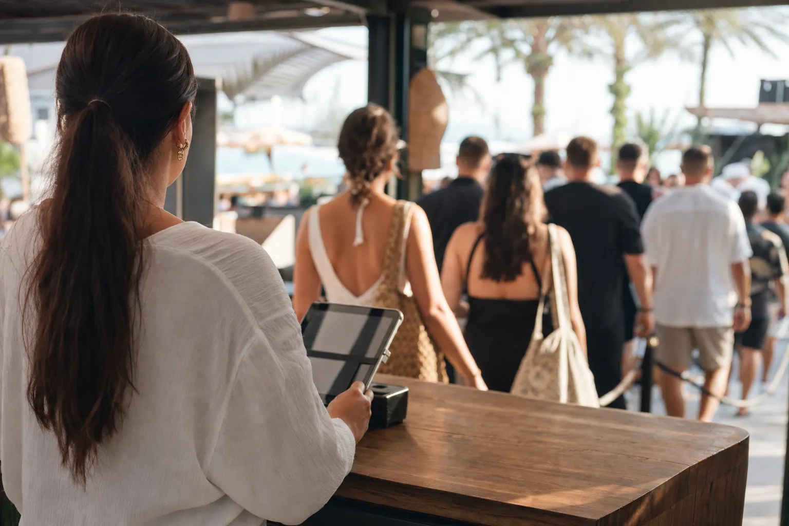 Host at a busy beach club entrance desk with a tablet, a queue of arriving guests softly out of focus behind
